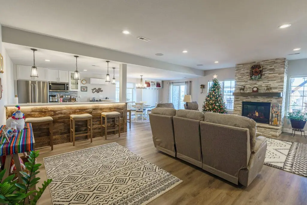 Open basement layout with a bar on one side, sectional seating and a stone fireplace with mounted TV